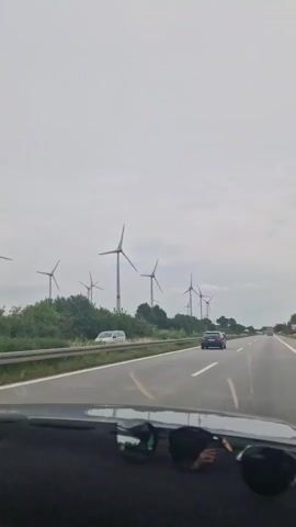 The video shows a view from inside a car looking out at a highway with a line of wind turbines on the left side of the road. The sky is overcast. There are several cars on the highway, including a dark-colored sedan driving ahead of the camera. Sunglasses are visible on the dashboard in the foreground.