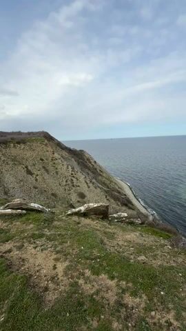 The video presents a static, wide shot of a coastal landscape on a cloudy day. The foreground features a grassy area, transitioning into a sloping, rocky cliff face covered in sparse vegetation. Several large pieces of driftwood lie scattered near the edge of the cliff.  The cliff leads down to a narrow, pebble beach that meets the water. The sea, which occupies the majority of the frame, stretches to the horizon under a sky filled with scattered clouds. The overall scene evokes a sense of natural, slightly rugged beauty.