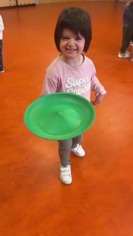A young girl in a pink sweater and grey pants is attempting to spin a green plate on a wooden stick in a room with an orange floor. She smiles at the camera initially. Other children are present in the background. The girl spins the plate on the stick, but it falls to the floor. There is a red curtain and wooden bench in the background.