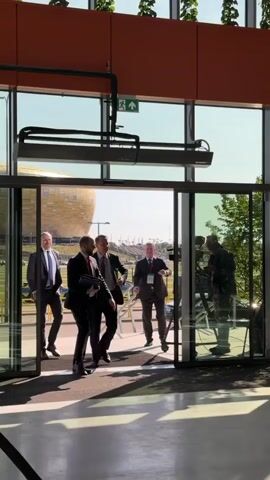 The video begins with three men in suits walking through large glass doors. One man on the far left is walking away from the camera. The second man from the left is walking towards the camera and holding a black folder. The third man from the left is also walking towards the camera and appears to be talking to the second man. There is a cameraman standing to the right of the glass doors, filming the group as they enter. As the video progresses, the first two men continue to walk through the doors. The third man is briefly visible talking to a woman in a black suit with a lanyard around her neck. The video then cuts to a shot of the two men walking away from the camera, exiting through another set of glass doors. A sign is visible on a pillar to the right that reads "UkraineRecovery CONFERENCE" and "21-22 June 2023 - Gdansk, Poland." The environment appears to be a modern building with large windows, and an outdoor area with trees and a stadium-like structure in the background. The lighting suggests it is daytime.