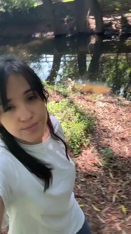 A woman with long, dark hair, wearing a white t-shirt, walks along a path in a wooded area. The path is covered in leaves. Trees with dense green foliage surround the path. Light filters through the leaves. A body of water, possibly a stream or pond, is visible at the beginning of the video. The woman appears to be filming herself.