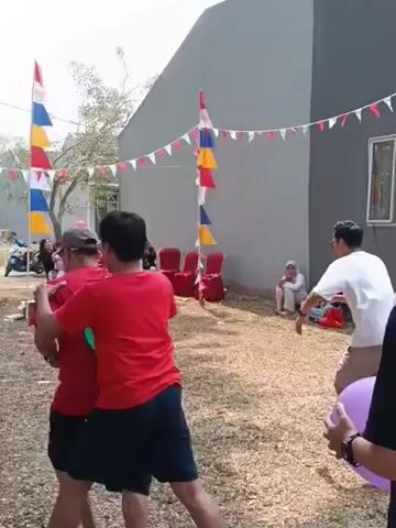 The video shows an outdoor event with several people participating in a game. Two groups of people are facing each other, suggesting a competition. One group, wearing red shirts and dark shorts, appears to be in the foreground, with their backs to the camera. They are holding what look like balloons or water balloons, indicating a game involving throwing or bursting them. The other group, wearing white shirts and shorts, is in the mid-ground, facing the first group. They seem to be defensively positioned, possibly trying to avoid being hit. The environment is a dry, grassy area with a building in the background. The building is decorated with flags and bunting, suggesting a festive occasion. Several onlookers are seated in chairs on the sides, observing the game. There are also children present, one of whom is holding a yellow object. The overall scene depicts a lively outdoor activity, likely a game of chance or skill involving water balloons.