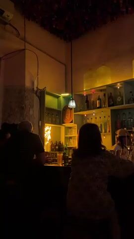 The video captures the interior of a dimly lit, cozy bar, likely during the evening. Warm, amber-toned lighting permeates the space, creating an inviting atmosphere.

Two patrons are seated at a wooden bar counter, their backs mostly to the camera. On the left, a bald man wears a light-colored, short-sleeved shirt with a tropical pattern featuring palm trees. On the right, a person with dark, long hair is dressed in a patterned shirt. Briefly, a bartender in a light-colored cap and dark shirt can be seen behind the bar, attending to drinks. Towards the end of the video, a hand holding a phone enters the frame on the far right, possibly taking a picture.

Behind the bar, a brightly lit shelving unit displays numerous liquor bottles, arranged on multiple tiers of teal or light blue shelves. Small, colorful string lights are draped along these shelves, adding to the festive mood. Above the shelves, large, green-tinted glass bell jars or demijohns are prominently displayed, lit from below. Other decorative elements include smaller green-tinted bottles, a yellow neon sign (text unclear), and a framed religious image, likely of the Virgin Mary, tucked into a corner shelf.

The ceiling appears adorned with dark red or patterned fabric streamers. Hanging pendant lights with green and white glass shades illuminate the bar counter. A light-colored tote bag is visible on the bar counter in front of the right-side patron. The eclectic decor, including the religious imagery and warm lighting, suggests a unique establishment, characteristic of many bars in Mérida, Mexico.