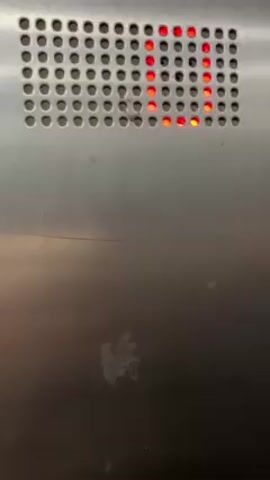 The video shows a close-up of a metal surface with a perforated panel containing red LED lights. The lights illuminate sequentially, spelling out the word "LOADING". The metal surface has a few scratches and a faint, faded mark or stain near the bottom. The environment appears to be indoors, possibly inside an elevator or similar enclosed space. No people are visible.