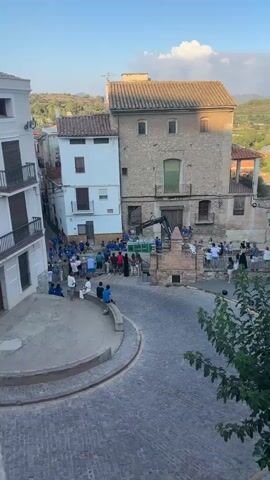 The video captures an outdoor scene in a village square. The time appears to be late afternoon or early evening, given the lighting. The square is paved with cobblestones and has a circular fountain or seating area in the foreground. In the background, there are several buildings, including a white building and a larger, older stone building with a tiled roof. There is also a crane or construction equipment positioned near the entrance of the stone building. A group of people, varying in age and gender, are gathered in the square. Some are standing, some are sitting on the fountain's edge, and others appear to be interacting with each other. Many are wearing blue shirts, suggesting a common affiliation or participation in an event. The overall impression is of a community gathering or event taking place in a historic village setting. The environment suggests a Mediterranean or similar climate, with vegetation visible in the background.