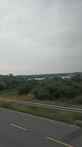The video shows a view from a moving vehicle, likely a car or bus, looking out a side window. The scene depicts a rural landscape under an overcast sky. A paved road with yellow lane markings runs across the bottom foreground of the image. To the left of the road, a metal guardrail separates the road from a grassy embankment. Beyond the embankment, a lush green field stretches out, bordered by a dense line of trees and vegetation. In the distance, a body of water, possibly a lake or river, is visible through the trees. The overall impression is one of a journey through a verdant, natural setting.