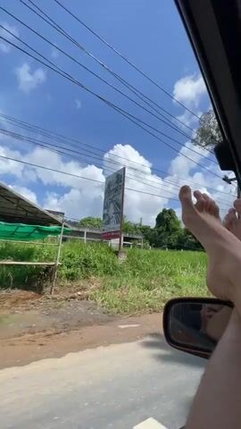 The video begins with a close-up of a bare foot resting on the exterior of a moving vehicle. The camera then pans to show lush green foliage and a bright blue sky with scattered clouds outside the car window. The perspective shifts to inside the car, revealing the driver's legs as they steer the vehicle. The dashboard, including the steering wheel and instrument cluster, is visible. The scene depicts a person driving a car through a scenic, rural landscape on a sunny day.