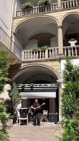 The video shows a courtyard with an arched colonnade. In the foreground, a man in a black shirt and pants plays an acoustic guitar. He is standing near a stage setup with speakers. Above him, on the second-floor balcony, a person in a white top stands facing away from the camera, holding their hair. Another person in a blue shirt is visible below them on the same balcony. The architecture suggests a European setting with stone and stucco elements. There are plants visible in pots on the balconies and in the courtyard. The lighting suggests it is daytime with sunlight casting shadows.