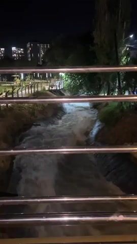 The video shows a body of water flowing through a man-made channel at night. The water is churning and appears to be moving quickly, with some turbulence visible. The channel is lined with dirt and vegetation on both sides. A metal railing, consisting of horizontal bars, is in the foreground, partially obscuring the view of the water. In the background, trees are visible, and beyond them, buildings are illuminated. Streetlights are also present, casting a glow on the scene. The overall impression is of an urban or suburban setting at night with a flowing water feature. There are no people visible in the frame.