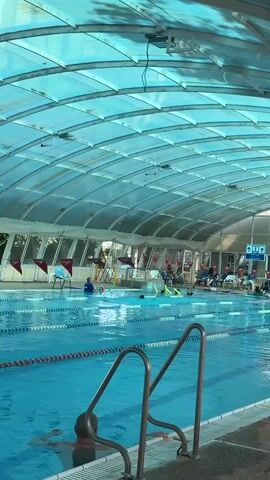 The video shows an indoor swimming pool with a translucent, curved roof. There are multiple swimming lanes marked with ropes. Several people are in the pool, some swimming and others standing. A few individuals are sitting in chairs on the pool deck, and there is a life preserver attached to a pole near the right side of the frame. The overall environment appears to be a recreational or public swimming facility.