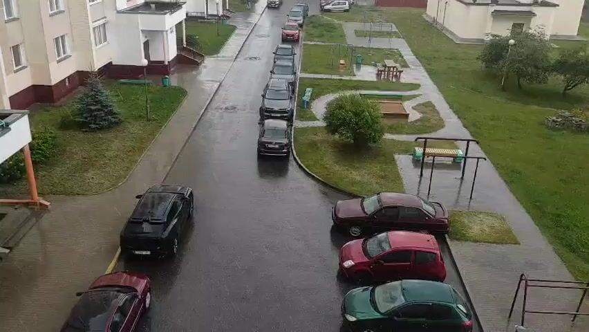 The video shows a rain-slicked street with parked cars and apartment buildings. A line of cars is parked on the street. A black SUV is parked on the left side of the street. In the foreground, a burgundy sedan is parked on the left, and a green hatchback is parked on the right. In the background, apartment buildings line the street. A playground with a sandbox and picnic tables is visible on the right. The sky is overcast, suggesting a rainy day.