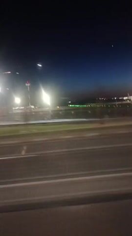 The video depicts a nighttime view from a moving vehicle, likely a car, showing a road and illuminated structures or lights in the distance. The road is asphalt with lane markings, and the adjacent area features a grassy section with bright lights. The background reveals a dark sky with distant lights, suggesting an urban or developed area. The camera movement indicates travel along the road. There are no people visible in the scene. The video appears to be captured through a slightly dirty or foggy window, affecting the clarity of the image.