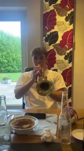 A young man with short curly hair, wearing a white t-shirt, sits at a wooden table and plays a brass trumpet. The table is cluttered with a half-eaten bowl of food, empty Corona beer bottles, a phone, a spoon, and a Corona box. A large window behind him shows a green lawn with trees and shrubs. To his right, a wall is decorated with a colorful floral pattern. He is centered in the frame, facing forward, focused on playing the instrument.