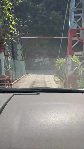 The video begins from the interior of a vehicle, showing a wooden bridge ahead. A red and white barrier arm is visible. The vehicle approaches the bridge and crosses it. The landscape consists of a dense forest on either side of the road. The bridge appears to be narrow and made of rough wooden planks. As the vehicle moves forward, the barrier arm is lifted or already up. The video ends as the vehicle continues past the bridge and barrier.