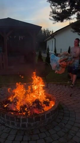 A man is seen standing next to a large bonfire in a backyard. He is shirtless and wearing swim trunks, holding a bundle of trash, which appears to be plastic bags and paper, over the flames. The bonfire is contained within a circular brick fire pit in the center of a paved patio. In the background, there is a wooden gazebo on the left and a white house with a dark roof on the right. The sky is a dusky twilight hue, suggesting it is either early evening or late dusk. The overall mood seems casual, as the man is adding refuse to the fire, perhaps for disposal. There are no visible text elements in the video.