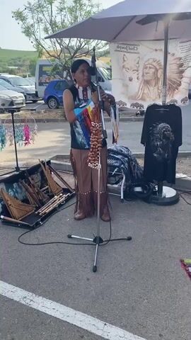 A male performer dressed in a traditional Native American outfit plays a flute. He wears a colorful fringed vest, a beaded necklace, and brown pants with fringe. He stands in front of a microphone on a stand. To his left is an open case containing several flutes. To his right, a display board shows a t-shirt with a Native American design. A large umbrella is set up behind the performer, providing shade. In the background, a parking lot is visible with several cars and vans. A red rug is spread on the ground in front of the performer, displaying various handmade items, including dreamcatchers, beaded necklaces, and more flutes. The scene appears to be an outdoor market or festival.