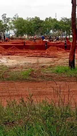 A time-lapse video shows a construction site from a slight distance, with green grass in the foreground and a line of trees in the background under an overcast sky. The ground is reddish-brown dirt, with several excavated trenches and piles of dirt. Numerous workers, dressed in casual clothing, are busy moving dirt with shovels and wheelbarrows, and some are working in the trenches. A cement mixer is visible, along with bags of cement and piles of gravel. A tall, slender dead tree is in the foreground on the left. The time-lapse effect makes the workers appear to move quickly, and the activity progresses from the initial excavation to the laying of foundations.