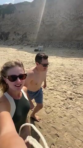 A woman and a man are walking on a sandy beach. The woman is in the foreground, smiling at the camera, and holding the camera. She is wearing sunglasses and a swimsuit. The man is behind her, also looking towards the camera, and smiling. He is wearing shorts. The beach appears to be next to a cliff. The wind is blowing the woman's hair.