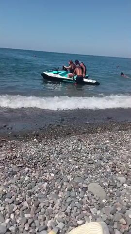 A group of people are interacting with a jet ski on a pebbly beach. The jet ski is partially in the water, with three people on it. Two individuals are standing and appear to be pushing or adjusting the jet ski, while a third person is seated. In the background, several other individuals are in the water, swimming or wading. The water is blue and calm, with small waves breaking on the shore. The sky is clear and blue, indicating a sunny day. The beach is covered in a variety of small, smooth pebbles. A person's feet are visible in the foreground, suggesting the video is filmed from a seated perspective on the beach.