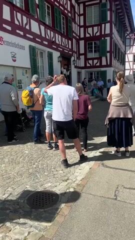 A group of people are standing in front of a building, watching a man in costume standing in the doorway. The building is a white and red half-timbered building. The man in costume is wearing a black hat and a white shirt with a black vest. He is holding a sword in his right hand and a staff in his left hand. He is speaking to the group of people. Some of the people are standing and some are sitting on steps. The ground is paved with cobblestones. There is a drain in the ground. The sky is sunny.