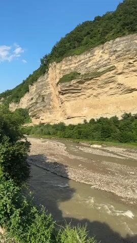 The video shows a wide, shallow river with a rocky bed flowing through a valley. The riverbanks are lined with lush green trees. A steep, rocky cliff face rises on the left side of the valley, covered in vegetation. The sky is a clear blue with a few wispy clouds. The scene appears to be a natural landscape, likely in a mountainous or hilly region, and the lighting suggests it is daytime. There are no people or man-made objects visible in the frame.