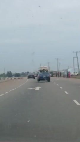 The video, shot from inside a moving car, shows a highway road with multiple lanes separated by painted lines. In the center of the frame, another car, a blue sedan, is driving ahead. It is carrying large bundles tied to its roof rack. Other vehicles are visible in the distance, and the shoulder of the road is lined with concrete barriers. Power lines are visible on the roadside. The sky overhead is overcast, and the road surface appears wet. The focus of the camera shifts as the video progresses, sometimes blurring the view.