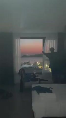 The video shows a man in a hotel room standing by a window with a beautiful sunset view. The room appears slightly messy, with clothes strewn on a chair and bed. The man is wearing a hat, a dark t-shirt, and shorts. He picks up a mobile phone from the windowsill and looks at it. The background showcases a coastal view with lights and a colorful sky.