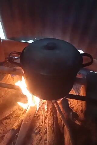 A black pot with handles sits on a rustic, makeshift stove constructed from logs and possibly rocks. The stove is fueled by a visible fire, with flames licking the bottom of the pot. The surrounding environment appears to be outdoors or in a dimly lit, primitive structure, suggested by the rough wooden supports and a dark, corrugated background that could be a wall or roofing. The lighting is low, with the fire providing the primary illumination, casting a warm, flickering glow on the pot and its surroundings. The overall scene depicts a cooking setup using traditional or basic methods.