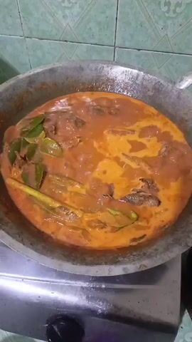 A cooking pot filled with a reddish-orange curry is being heated on a stove. The curry contains visible pieces of fish, several lime leaves, and stalks of lemongrass. The pot is a large, round, shallow metal pan with two handles on opposite sides, and it sits on a silver stove with a black knob visible on the front. The background shows a tiled wall with a green and white geometric pattern. The camera is positioned directly above the pot, looking down.