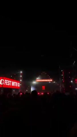 The video shows a crowd of people at a concert or festival at night. The stage is brightly lit with red lights and displays the text "FEST WITH COCA-COLA 20." The crowd is mostly silhouetted against the lights, some people are raising their hands in the air. The environment is dark, likely an outdoor venue, with lighting structures and screens visible around the stage. The atmosphere appears lively and energetic.