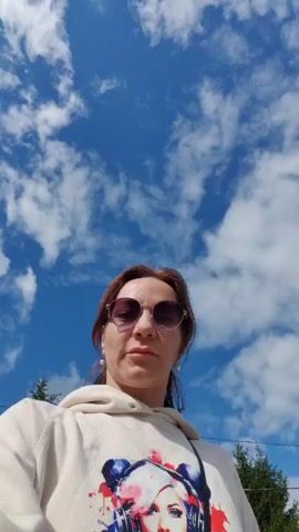 This media file features a woman in sunglasses and a light-colored hoodie with a graphic print of a woman's face wearing headphones. The scene appears to be outdoors during the day, with a bright blue sky and scattered white clouds visible in some shots. The woman is walking, and the camera captures her from various angles, including close-ups and wider shots showing her surroundings.

In one shot, the woman is walking on a dirt path, with her shadow cast on the ground. Another shot shows her walking alongside another person, and a few cars are visible in the background, suggesting a rural or suburban setting. Trees and houses are also present, contributing to the outdoor environment. The weather seems pleasant, indicated by the clear sky and the subjects not wearing heavy clothing. The overall mood appears casual and relaxed, as the woman is engaging in an activity like walking. The location is identified as Orekhovo, Russia. There is no visible text in the media file.