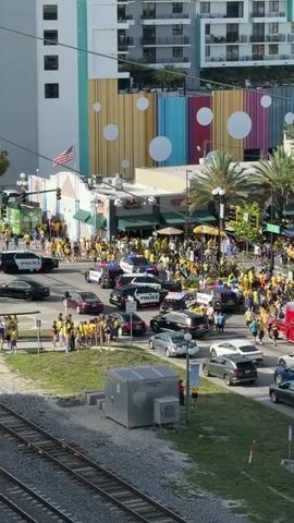 The video shows a crowded street scene with numerous vehicles, including police cars and an ambulance, suggesting a potential incident or event. People, many wearing yellow, are gathered on sidewalks and in the street, creating a lively atmosphere. Buildings lining the street display colorful murals and modern architecture. The presence of palm trees and a sunny sky indicates a warm, possibly tropical, location. The video pans across the scene, capturing different angles of the activity and the urban environment.