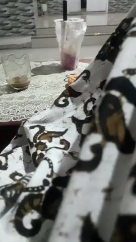 The video displays a close-up of a batik fabric with a white background and black and brown abstract patterns. In the background, a table is visible, set with a glass cup containing a small amount of brown liquid, likely coffee or tea. To the right of the cup, a taller plastic cup with a straw holds a pink beverage. A black lid is also present on the table. The scene appears to be indoors, possibly in a dining or living area, given the presence of furniture and what looks like a tiled floor or wall in the background. The lighting suggests it might be daytime or early evening, as it's not overly bright. The overall mood is relaxed and casual, with the focus on the textured fabric and the beverages. The setting and the batik pattern hint at a cultural context, possibly Indonesia. No people are visible in the frame, and there is no discernible text in the video.