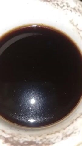 A close-up shot shows a white ceramic mug filled with a dark liquid, likely coffee or tea. Tiny bubbles are visible on the surface of the liquid, and a bright reflection, possibly from a light source, is prominent in the lower portion of the mug. The textured rim of the mug is also visible. The scene is static, with no discernible action or movement.