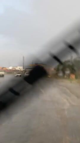 A wet road is visible with several cars driving on it. The sky is overcast and it appears to be raining or has recently rained. There are some buildings and industrial equipment in the background.