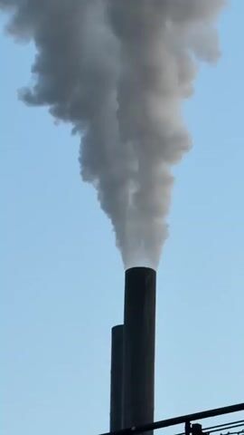 The video shows two industrial smokestacks against a clear blue sky. Smoke billows from the top of the taller smokestack, forming a large, grey cloud that drifts upwards and to the left. A second, shorter smokestack is visible next to the first, but no smoke is visibly emerging from it. The bottom portion of the image shows a dark, horizontal structure, possibly part of the industrial facility. The perspective is looking upwards at the smokestacks.