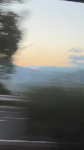 The video shows a view from a moving vehicle looking out at a landscape. The sky is a soft gradient of light blue and orange, with a few scattered clouds. In the distance, there are rolling hills or mountains that appear hazy and muted in color. In the foreground, there is a blurry expanse of green, likely fields, and dark areas that could be trees or shadows. The motion blur indicates that the perspective is shifting rapidly. There are no people or identifiable objects visible beyond the landscape itself.