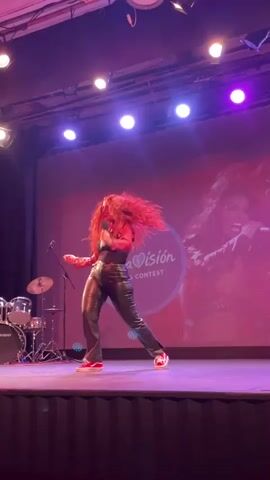 A woman with long, curly red hair stands on a stage, holding a microphone and singing. She is wearing a red leather jacket, a black top, and shiny black pants. Her shoes are red and white sneakers. Behind her, a large screen displays the words "AulaVisión Song Contest" and an image of a woman singing. There is a drum set to the left of the woman on stage. The stage is lit with purple lights.