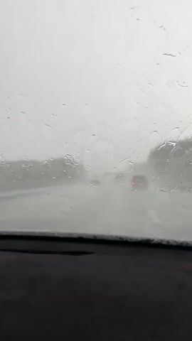 The video shows a view through a car windshield during heavy rain. Water droplets streak across the glass, obscuring the view of the road ahead. Cars with their brake lights illuminated are visible in the distance, driving through the downpour. The wipers are intermittently clearing a portion of the windshield, offering brief glimpses of the rainy conditions. The overall atmosphere is gray and hazy due to the weather.