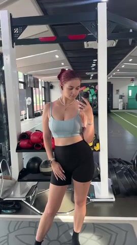 A young woman with red hair stands in a gym, taking a selfie in a mirror. She is wearing a light blue sports bra and black biker shorts. She adjusts her hair and touches her stomach as she poses. In the background, gym equipment, including weights and boxing gloves, is visible. The gym has a track with green turf.