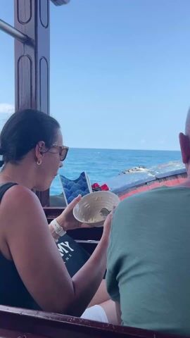 The video shows the inside of a boat on the water on a clear day. A woman in sunglasses is sitting and eating from a bowl. An older man in a green shirt sits next to her, his back to the camera. The camera pans to reveal two more men eating from bowls. One is shirtless with a beard and is sitting on the bow of the boat. The other wears a white T-shirt and a baseball cap. The ocean and land are visible in the background.