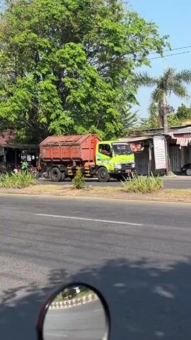 This media file depicts a bright, sunny daytime scene on a paved road, likely in Ketapang, Indonesia, given the location context. The primary subject is a bright green truck with a reddish-brown cargo bed, appearing to be a dump truck or garbage truck, driving on the right side of the road. The truck has a driver and a passenger visible in the cab, wearing what appears to be a green uniform.

The setting includes lush green trees, one particularly large one dominating the left side of the frame, and a palm tree on the right. There are buildings visible behind the truck, including a structure with a corrugated metal roof and what looks like a small shop or market on the left, with people gathered outside. Another building with a sign is present on the right. The road itself is asphalt, with a yellow line marking the lanes.

The activity taking place is simply the truck driving down the road. The overall mood is mundane, a snapshot of everyday life. The weather appears to be warm and clear, suggested by the bright sunlight and shadows. The time of day is clearly daytime, likely mid-morning or afternoon, based on the angle of the sun. The reflection in the bottom of the frame, showing a portion of a motorcycle's rearview mirror and a road, indicates the recording was likely made from a moving motorcycle.

No discernible text is visible or legible in the image.