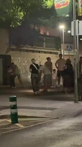 The video shows a group of people standing on a street at night, some of them near a building with a stone wall. There are a few small sparks or lights on the ground in front of some of the people. One person is bending down towards the sparks. The environment appears to be outdoors, possibly a town or city street, with some trees and streetlights visible.