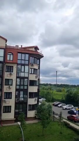 This media file presents a view of a residential area in Romashkovo, Russia, under a cloudy sky. The scene is dominated by a multi-story apartment building on the left, with a reddish-brown roof and numerous windows. The building appears to be modern, with visible air conditioning units.

In front of the building is a parking lot with several cars, including white SUVs, a red sedan, a black SUV, and a yellow car. A pedestrian walkway with a decorative fence borders the parking area. Green grass and trees are visible in the foreground and along the perimeter of the parking lot. Beyond the parking lot, a dense line of green trees stretches across the middle ground, with a tall, slender tower (likely a telecommunications tower) visible in the distance behind them. The horizon is dotted with buildings, suggesting the presence of a more urbanized area further away.

The weather is overcast, with heavy, grey clouds filling the sky. This suggests it might be a cool or potentially rainy day. The time of day is difficult to determine precisely due to the lack of direct sunlight, but it appears to be daytime.

No discernible human activity is taking place in the immediate visible area, apart from possibly a person near one of the cars. The overall impression is one of a quiet, suburban or semi-urban residential setting.

Visible text includes road signs with symbols and numbers, such as a "P" for parking, a pedestrian crossing symbol, and a speed limit sign indicating "20". A green garbage bin with some Cyrillic text is also visible.