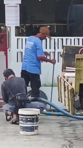 Two individuals are engaged in some activity outdoors, with one person standing and the other kneeling. The standing individual, wearing a blue shirt and a brown cap, holds a long pole or stick. The kneeling individual, dressed in a dark shirt and a dark cap, is focused on something on the ground in front of them. Between the two individuals is a white bucket with blue and black markings, and a thick, multi-colored hose. The background suggests an urban or semi-urban environment, possibly near a building with a white, slatted facade and a red structure on the left. The ground appears to be paved.