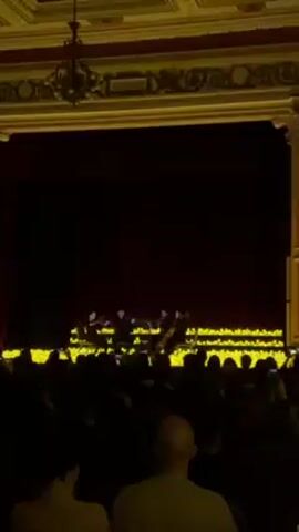 Here's a detailed description of the video:

The video shows a performance in a theater or concert hall. The stage is adorned with numerous small, warm-toned lights, creating a bright line just in front of a closed, dark-red curtain.  A group of musicians are partially visible on the stage in front of the curtain, seemingly performing.

The ornate architectural details of the building are visible, including decorative columns, balconies, and a chandelier. The walls are a warm, golden color.

An audience fills the seats, silhouetted against the light from the stage. Many individuals are holding up phones, likely to record the performance. The camera pans slightly to the left, revealing more of the auditorium and its architecture. The overall lighting in the auditorium is low, emphasizing the stage and the architectural details.
