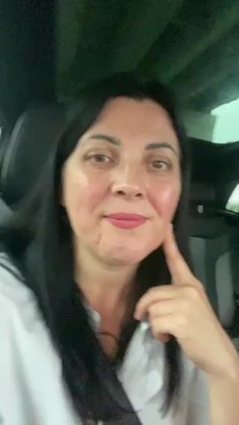A medium shot video shows a woman with long, dark hair and fair skin, wearing a white shirt and a black seatbelt, sitting in a car. Her left hand is near her face, touching her chin, as she looks at the camera. The background is blurred, showing the interior of a car. The lighting is dim, suggesting it is daytime or twilight.