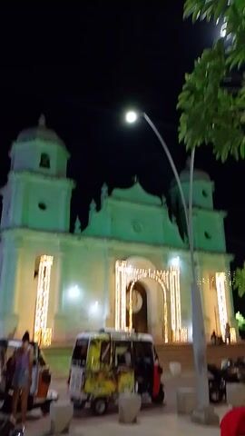 At night, a white church illuminated with lights stands tall. A large tree with green leaves is visible on the left, partially obscuring the view. A streetlight shines brightly near the tree. To the right, a bus or van with a red roof is parked. Several people can be seen near the church and on its steps. The overall scene is well-lit, highlighting the architectural details of the church.