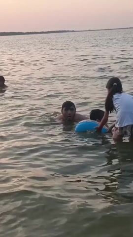 A group of people are swimming in a body of water, possibly a lake or the ocean, with a distant shoreline visible. The water is calm and appears to be shallow enough for people to stand in. In the foreground, an adult male is in the water with a young child wearing a blue inflatable ring around their waist. The adult is holding the child's arm. To the right, a young girl in a white shirt and shorts is also in the water, partially visible. In the background, several other individuals are swimming in a line, with their heads and shoulders above the water. The sky is a soft, muted color, suggesting either sunrise or sunset.