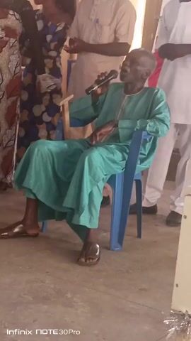 An elderly man is sitting in a blue plastic chair wearing a light green tunic and pants with leather sandals. He is holding a microphone to his mouth, appearing to be speaking.  Behind him, a woman in a patterned dress and two men in white shirts and pants are standing. The floor is a light-colored, smooth surface. The Infinix NOTE 30 Pro watermark is visible in the lower left corner of the frame.